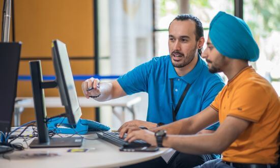 Two men working together at a computer workstation, one pointing at the screen while the other uses the keyboard.