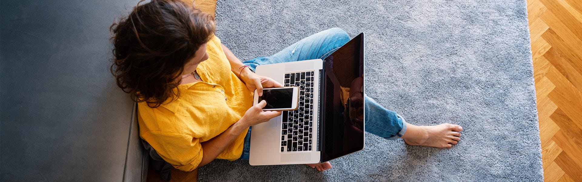 Woman sitting on loungeroom floor with laptop in lap and holding a mobile phone.