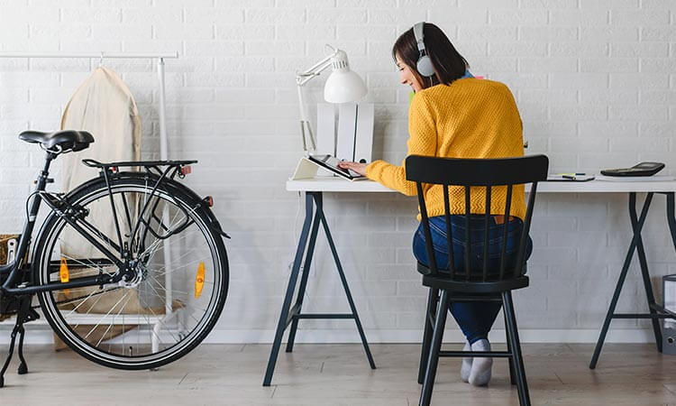 Student in yellow jumper and over-ear headphones sitting at desk in home office nook looking at tablet.