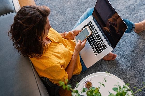Woman sitting on loungeroom floor, leaning against couch with laptop in lap and holding a mobile phone.