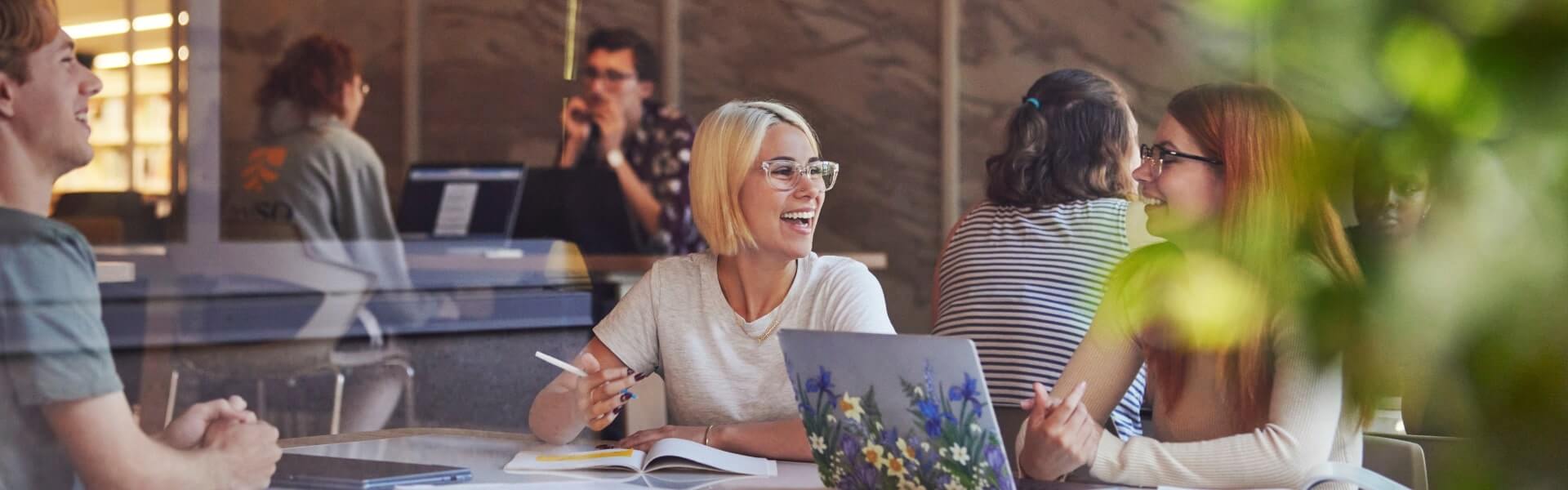 A group of people are sitting at a table in a cafe or library, working on laptops and notebooks, and having a conversation.
