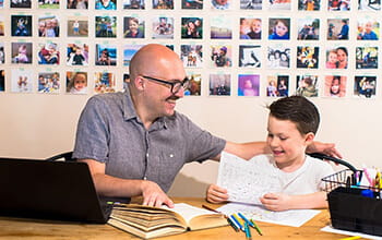 An adult and a child smiling and looking at a piece of paper at a desk with a laptop, books, and art supplies, with a wall of photos in the background.