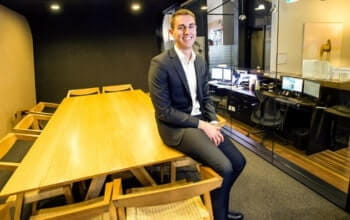 A young man in a suit sitting on a table in a modern office meeting room, smiling at the camera.