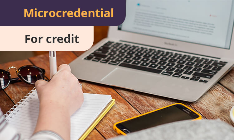 Person writing in a notebook at a wooden table with a laptop, smartphone, and sunglasses. Text on image reads "Microcredential For credit.