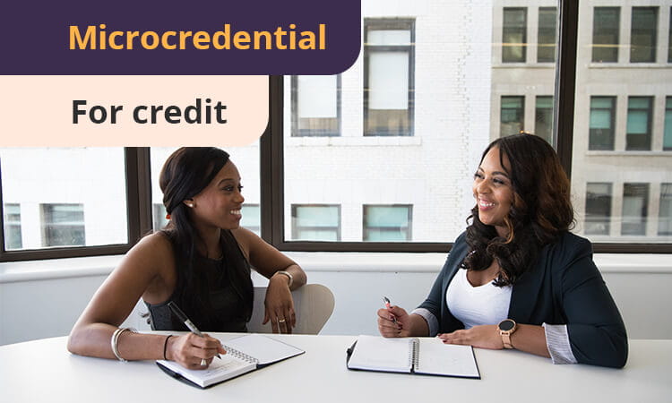 Two women are sitting at a table with notebooks, smiling and talking. A sign in the image reads "Microcredential For credit.