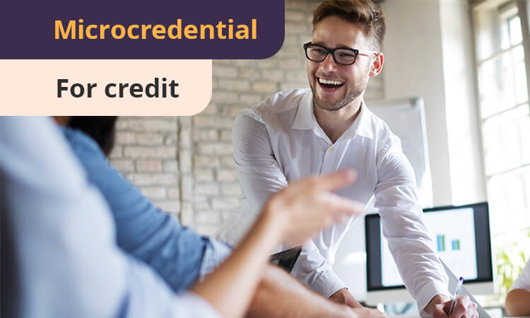 A smiling man in glasses leans over a desk, engaging with a seated person. The text overlay reads "Microcredential for credit.