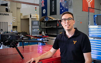 Adrian wearing glasses and a polo shirt with a logo smiles at the camera in a workshop environment, standing next to a drone with a toolbox and flags in the background.