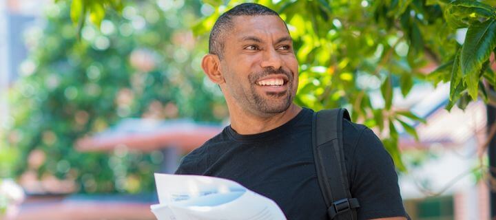 A smiling man holding papers and wearing a black shoulder bag outdoors.