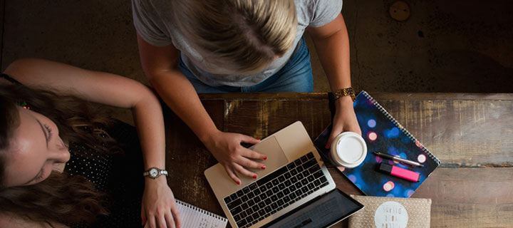 Birds eye view of two females, sitting at a timber table with laptops and study notes.