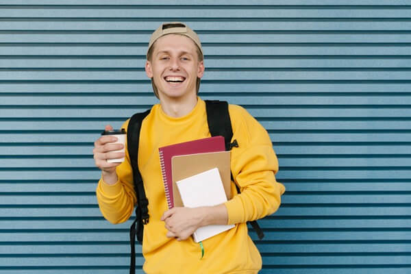 Young man in a yellow jumper, smiling at the camera and holding a coffee, books and backpack.