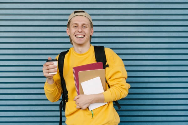 Young man in a yellow jumper, smiling at the camera and holding a coffee, books and backpack.