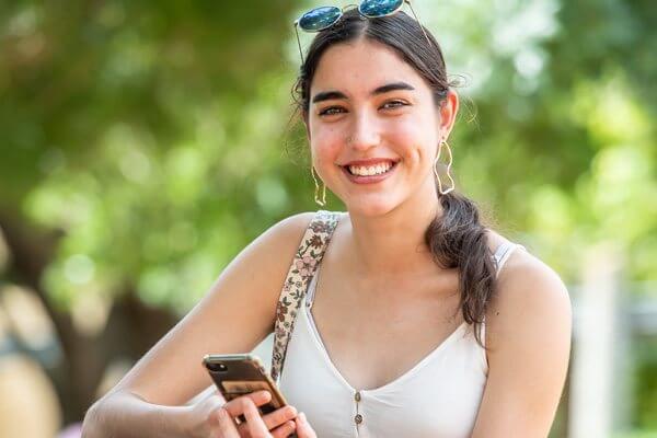 Young female sitting outside with her phone, smiling at the camera.