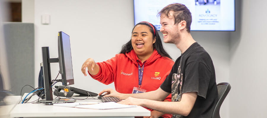 Two people sit at a desk working on a computer. One points at the monitor while both smile, suggesting a collaborative and positive interaction.