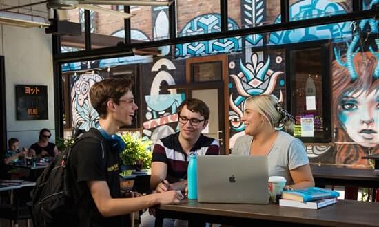 Three young adults sitting and chatting at a café with a computer and books.