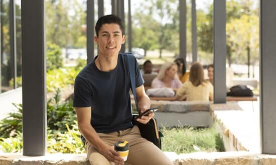 Young man sitting on a brick bench with a coffee and mobile phone, smiling at the camera.