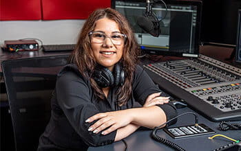 A smiling woman with headphones around her neck leaning on a radio mixing console in a studio.