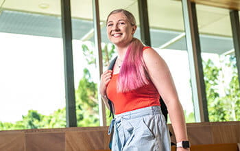 A smiling woman with pink-dyed hair carrying a backpack indoors.