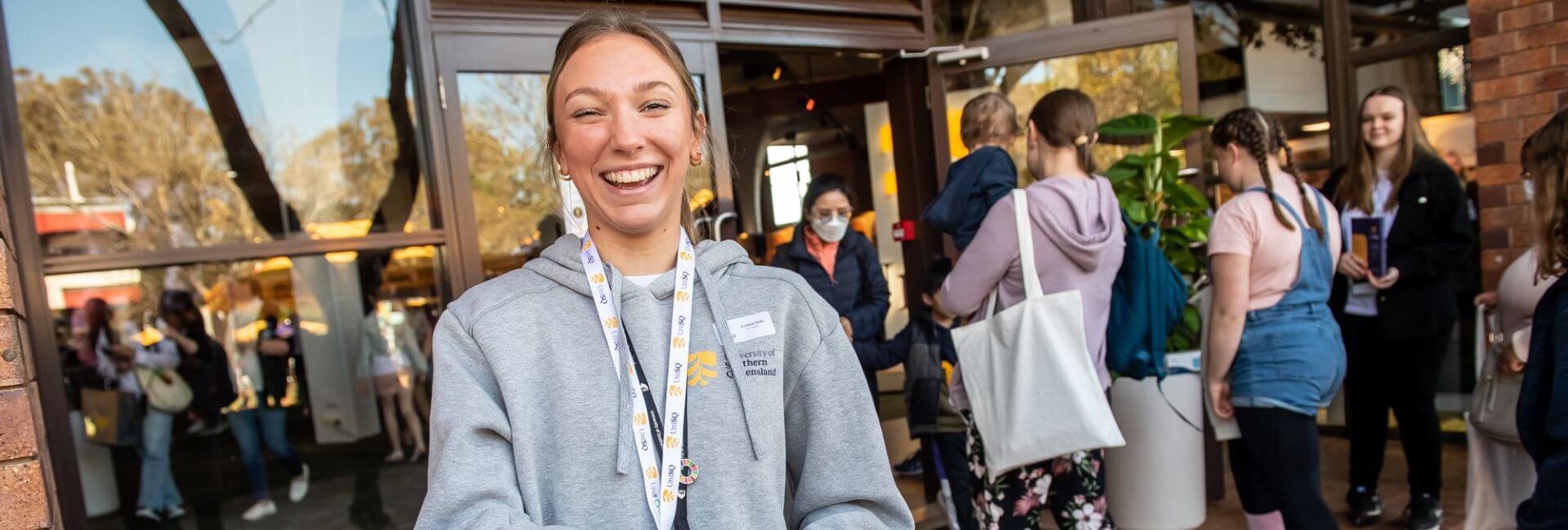 A smiling woman wearing a lanyard and hoodie greets at an event entrance, with other people in the background.
