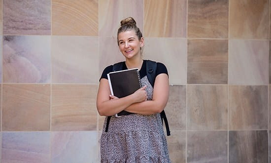 Student standing outside building holding study materials. 