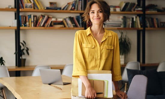 Young woman sitting on the edge of a timber table, holding a notepad and smiling at the camera.