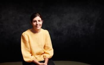 A woman wearing a yellow blouse is seated against a dark background and looking towards the camera.