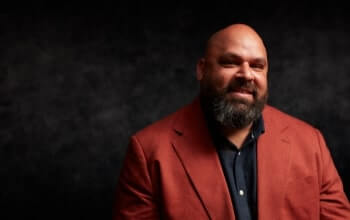 A bearded man in a red blazer smiles against a dark background.