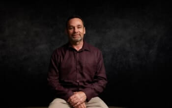 A man with short hair and a beard is seated against a dark background, wearing a maroon shirt and light-colored pants.