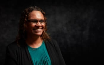 A lady wearing glasses, a teal shirt, and a black jacket is smiling against a dark background.