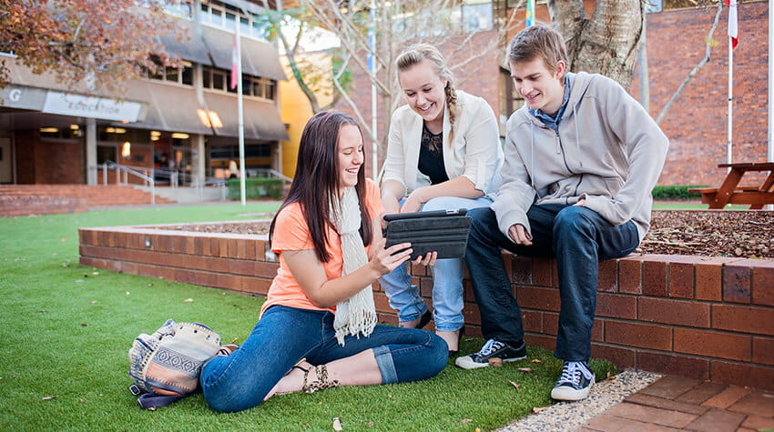 Three students using a tablet and smiling together the quad at UniSQ Toowoomba.