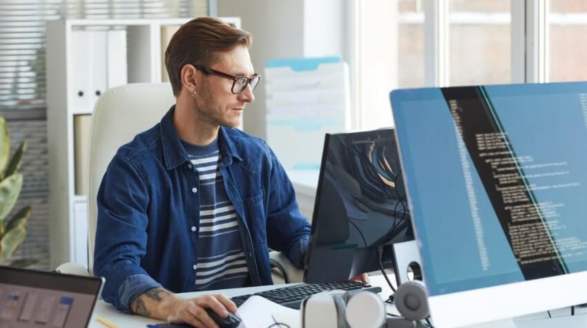 A man wearing glasses works at a computer with multiple monitors displaying lines of code in a bright office setting.