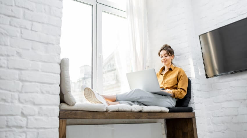 A woman sits on a window bench with her legs stretched out, working on a laptop in a bright room with white brick walls and a wall-mounted TV.
