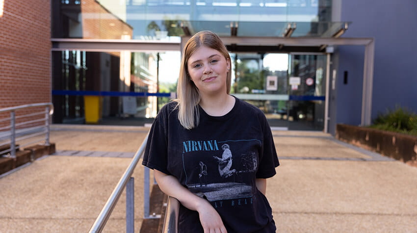 A young woman with blonde hair stands outside a modern building, leaning on a railing and wearing a black Nirvana T-shirt.