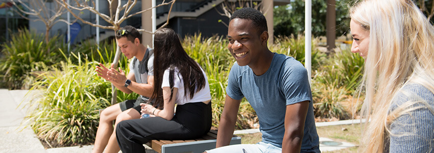 Four people are sitting and talking outside on a sunny day; two on a bench facing forward and two on a bench facing sideways.