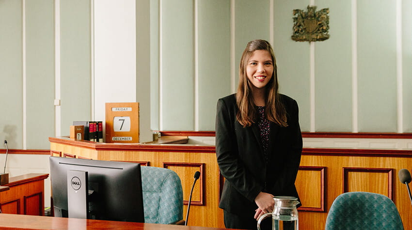 A young woman in business attire standing in a courtroom with a computer and water pitcher on the desk.