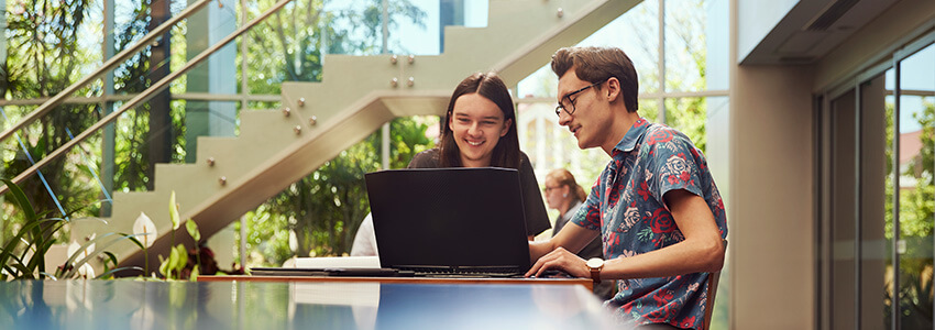 Two people sit at a table with a laptop, smiling. They are in a bright, modern space with plants and large windows.