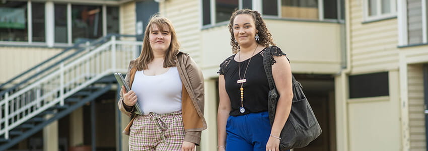 Two people walking outdoors, one holding a folder and wearing plaid shorts and a brown jacket, the other in blue pants and a black top, with buildings in the background.