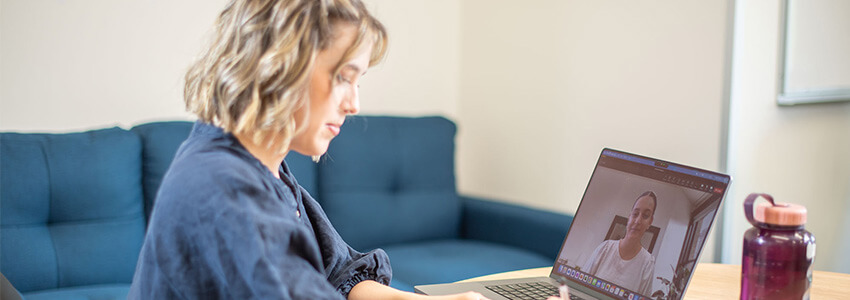 Woman sitting at a table, participating in a video call on a laptop, with a blue couch and a pink water bottle nearby.