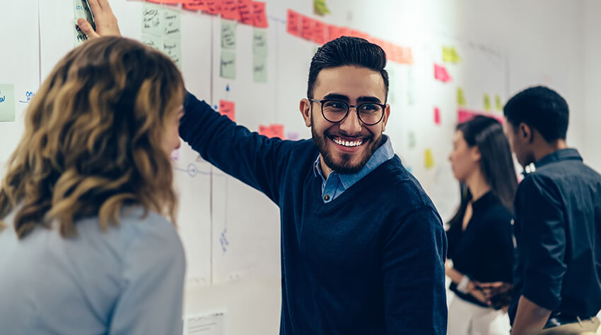 A man wearing glasses smiles while pointing at a whiteboard filled with notes, surrounded by colleagues engaged in discussion.