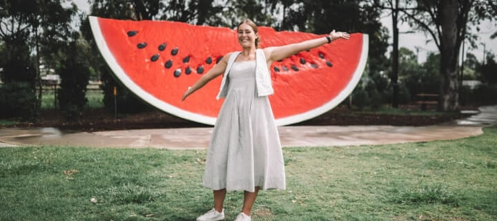 Woman smiling with arms outstretched in front of a large watermelon slice backdrop.