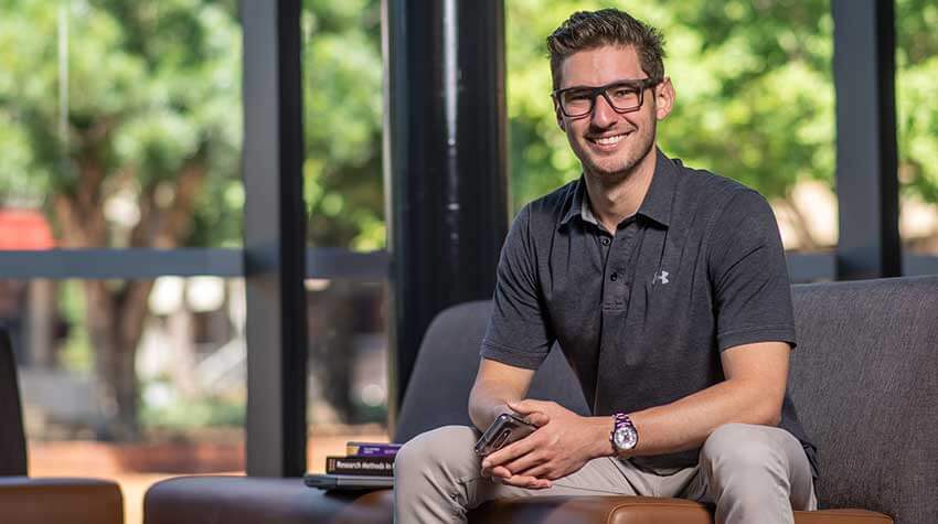 UniSQ student, Adam, sitting on a bench inside with textbooks by his side.