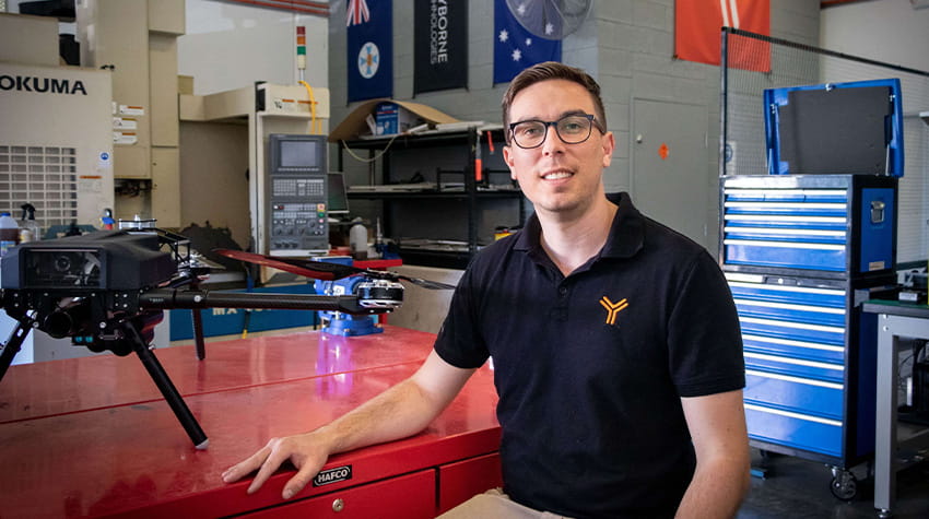 A man wearing glasses and a polo shirt with a logo, sitting at a red workbench in a workshop with a drone and flags in the background.