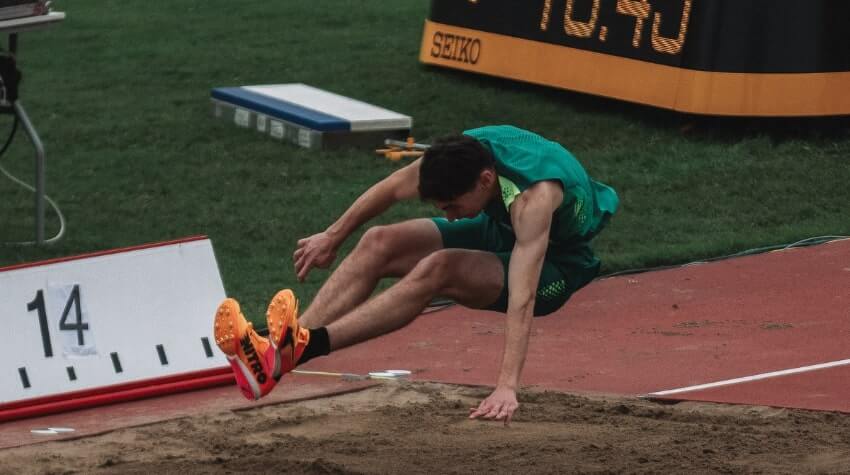 Athlete in green outfit lands in sand pit during long jump attempt at track and field event.