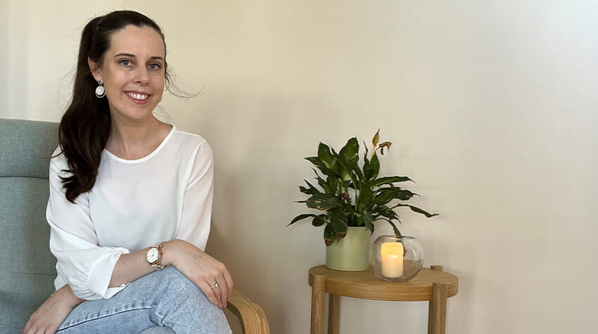 A woman smiling and sitting on a chair next to a small table with a plant and a lit candle.