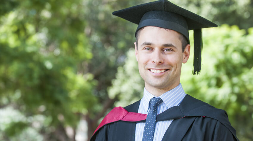 Andrew is wearing is graduation gown and mortar board, smiling at the camera.