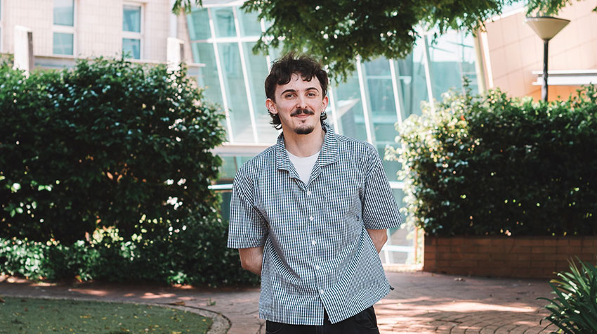 A man with short brown hair and a mustache stands outside in front of bushes and a modern glass building, hands behind his back.