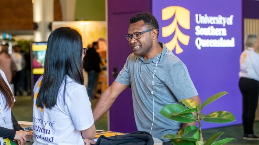 A man interacts with a woman at a University of Southern Queensland booth during an event. The booth has a purple backdrop with the university's logo.