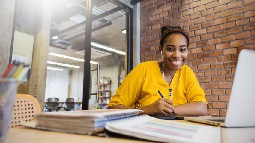 A smiling woman in a yellow shirt working at a desk with a laptop and notebooks in a brick-walled office space.