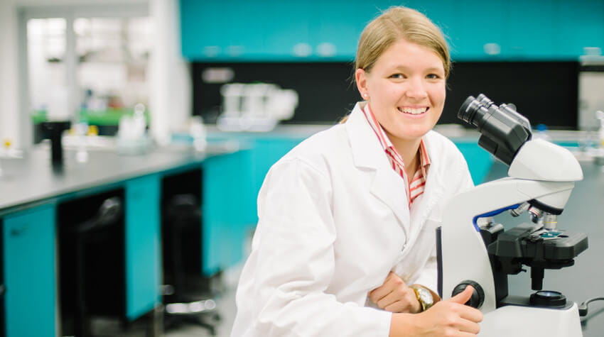 Constance is sitting in a laboratory with a microscope, smiling at the camera.