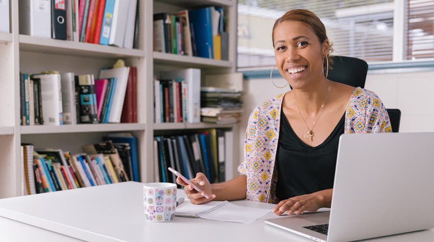 Despena is sitting at a desk, working on her laptop, smiling at the camera.