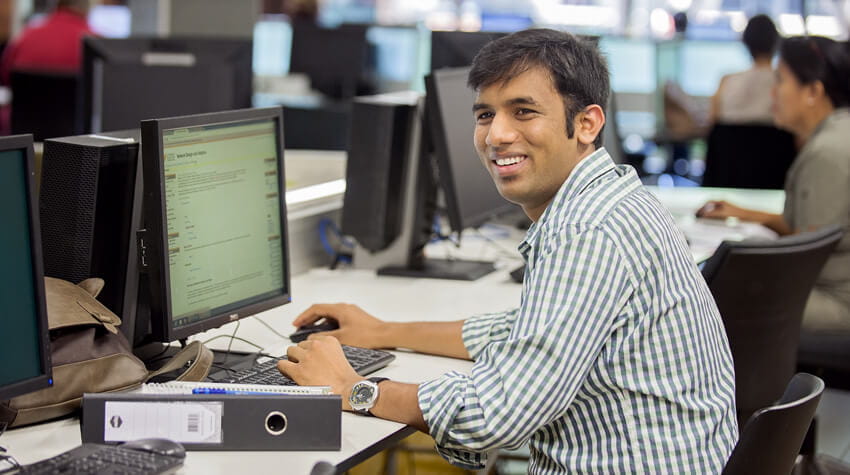 Dheerah is sitting at desk, working on a computer, smiling.